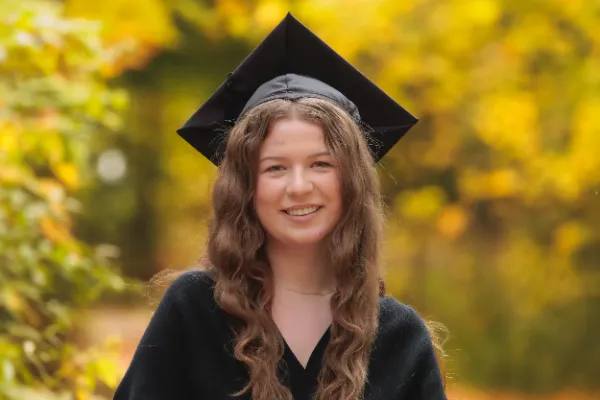 Graduation portrait of a woman in a black dress and mortarboard, smiling, with autumn foliage