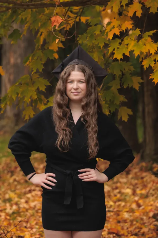 Grad girl black wrap dress mortarboard hands on hips autumn path