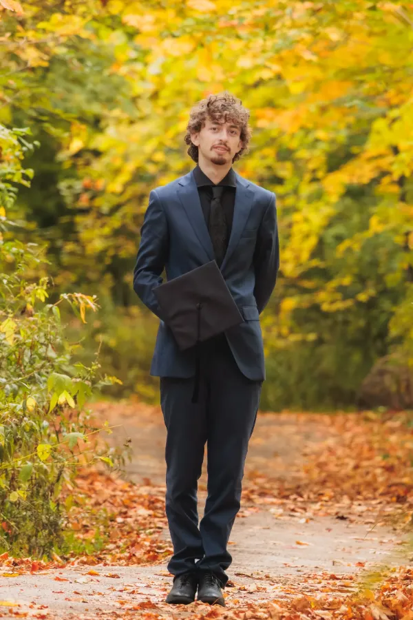 Grad guy blue suit holding mortarboard at side full body autumn path