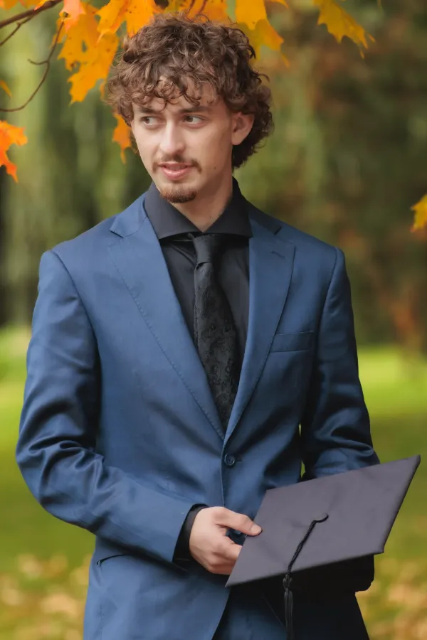 Grad guy blue suit holding mortarboard looking aside red maple