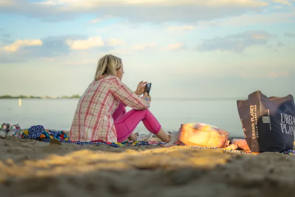 Woman pink outfit sitting beach sand looking at phone lake sunset