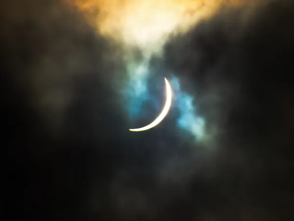 Solar eclipse thin crescent sliver through dark clouds blue teal glow